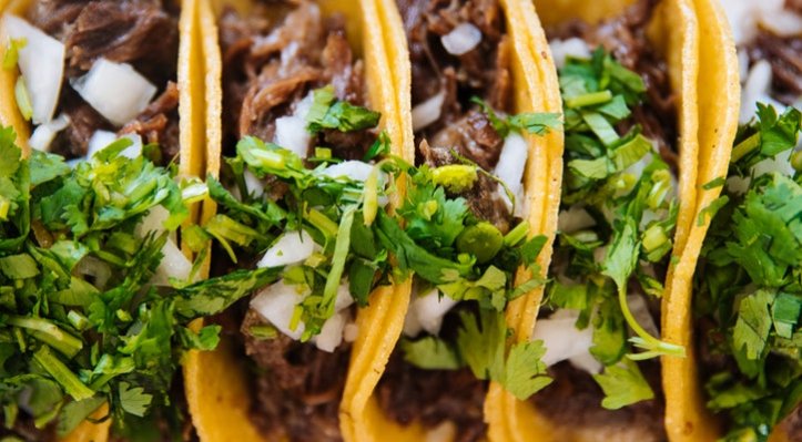 5 tacos in a row made of yellow corn tortillas, filled with steak, topped with white onions and cilantro